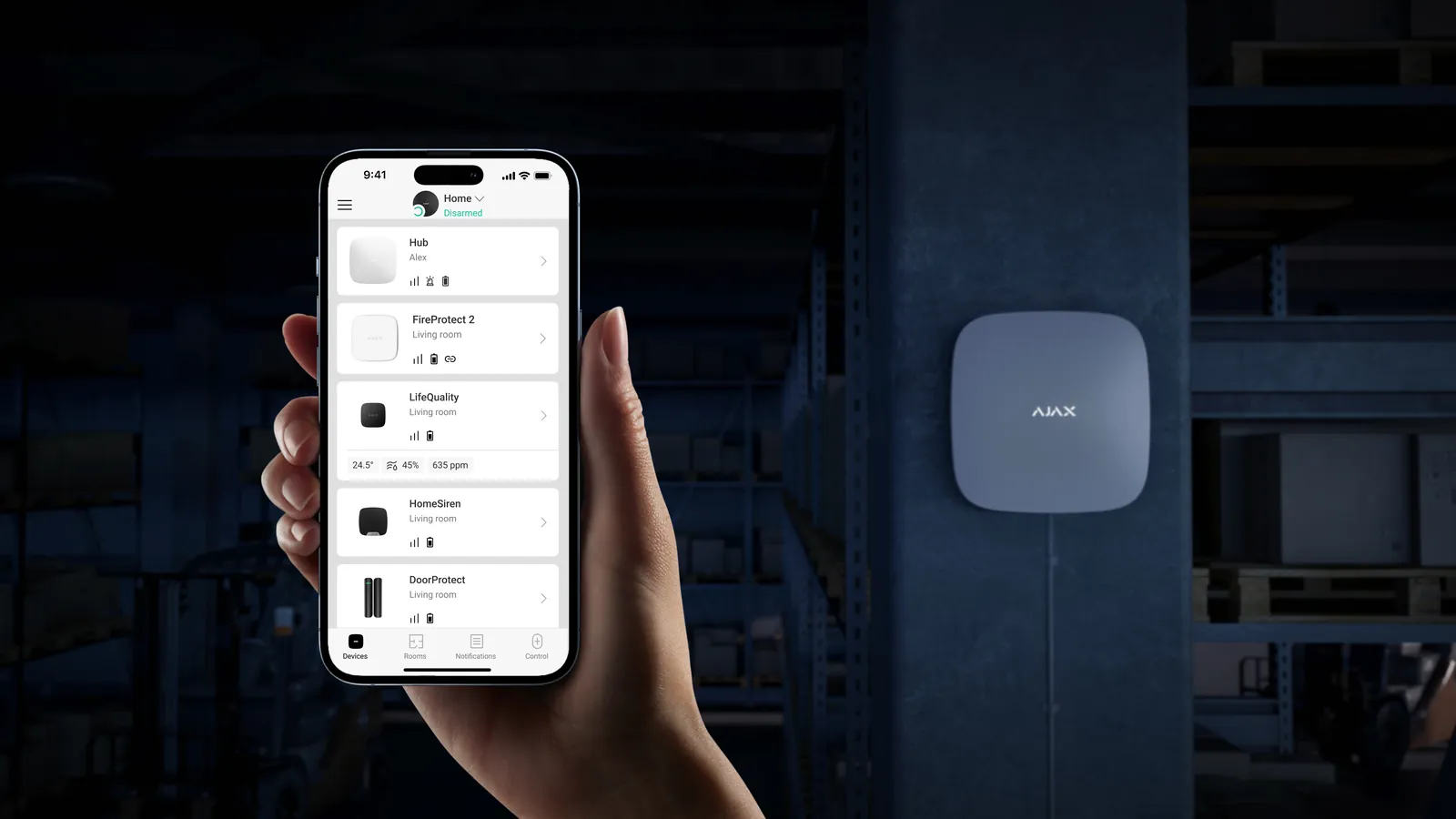 Hand met een smartphone met de Ajax-app, voor een zwarte Ajax Hub-centrale op een donkere muur