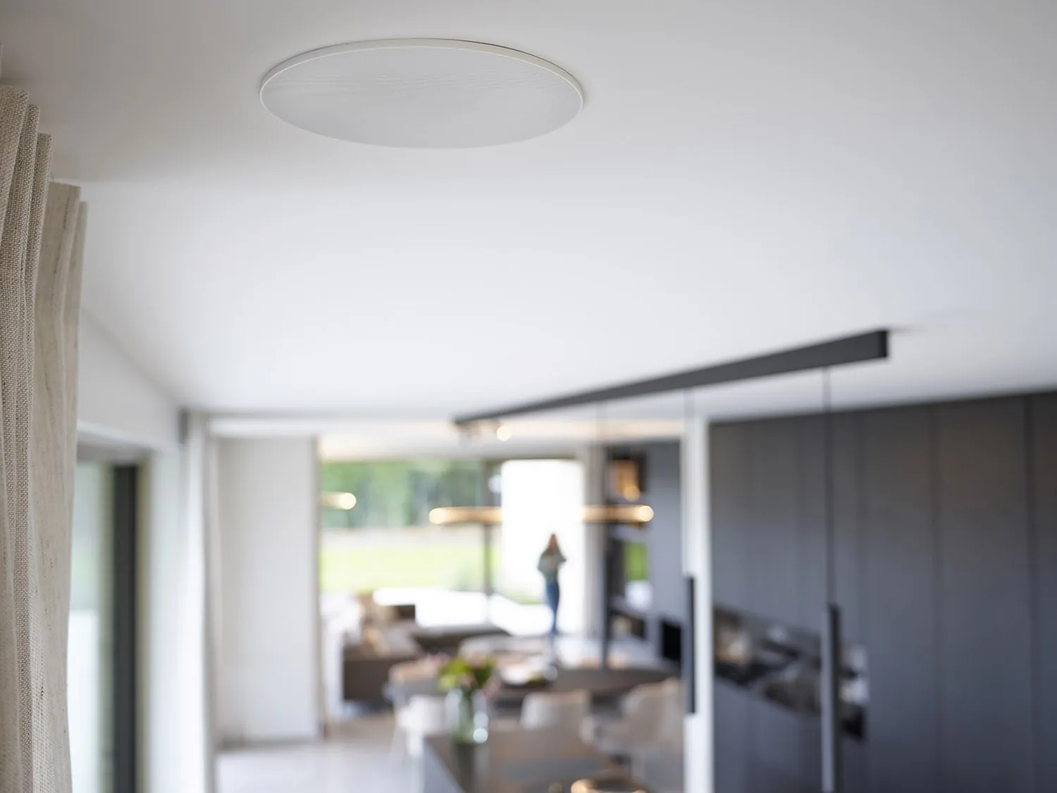 Enceinte ArtSound encastrée au plafond d’une maison, cuisine noire et séjour en arrière-plan