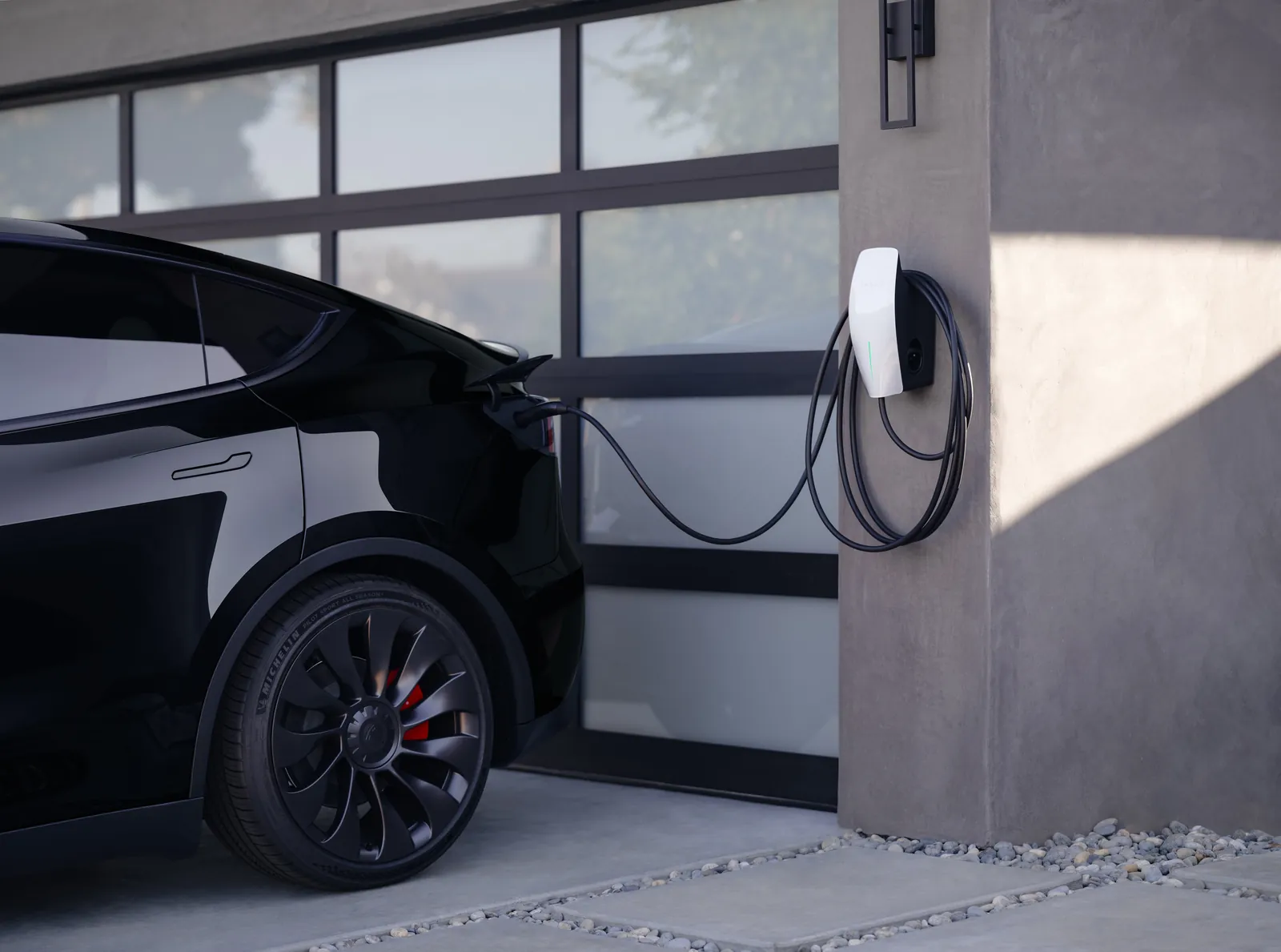 Tesla Universal Wall Connector on a grey wall beside a glazed garage door, with a black Tesla Model Y charging