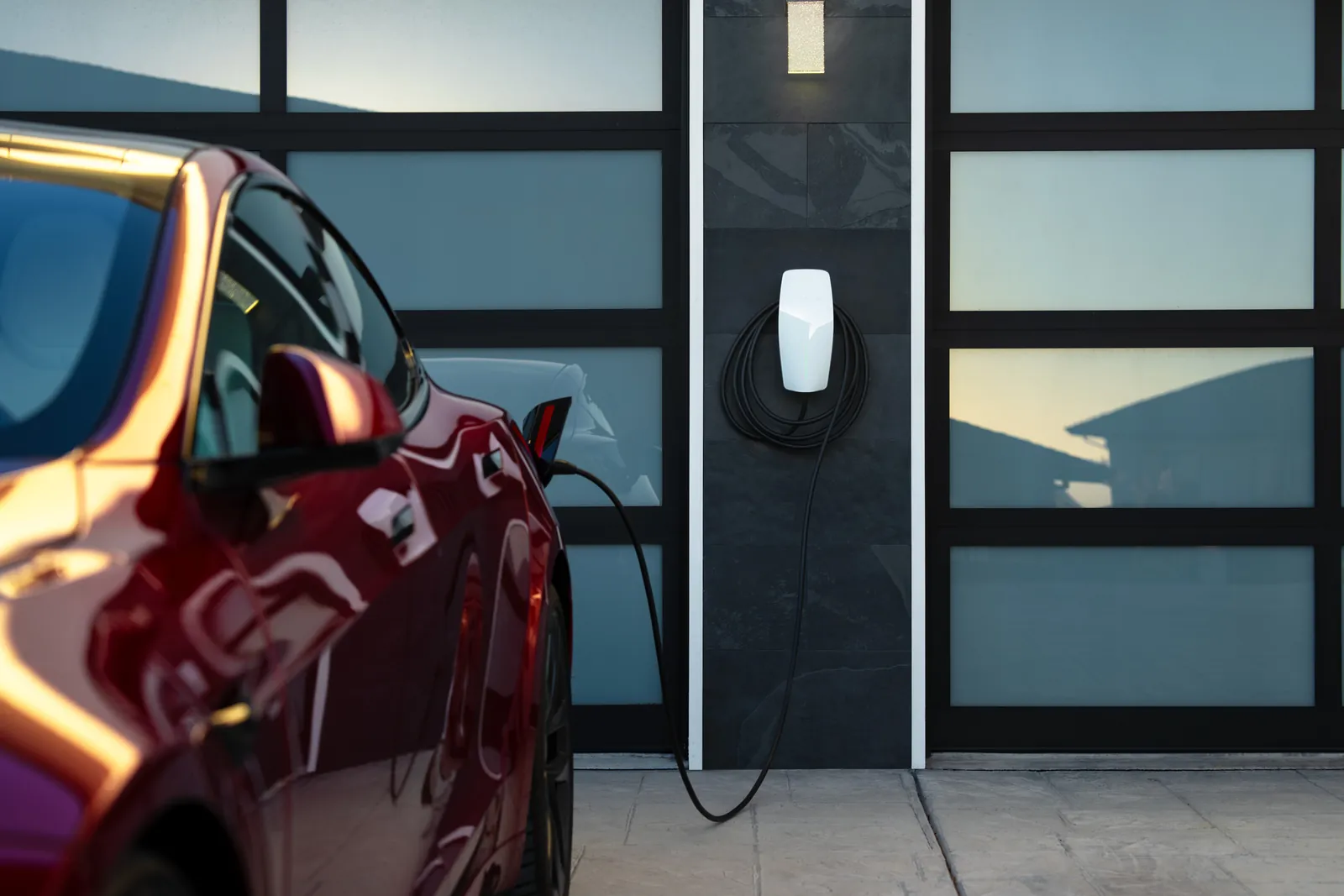 Tesla Wall Connector on a dark façade beside a glazed bay, with a red Tesla Model 3 charging